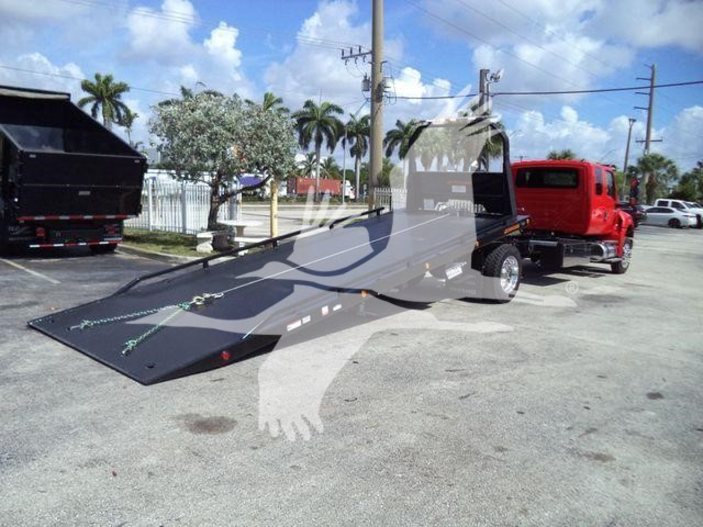 Flatbed rollback tow truck with a long black loading surface attached to a red truck, in a parking lot with palm trees and other vehicles in background.