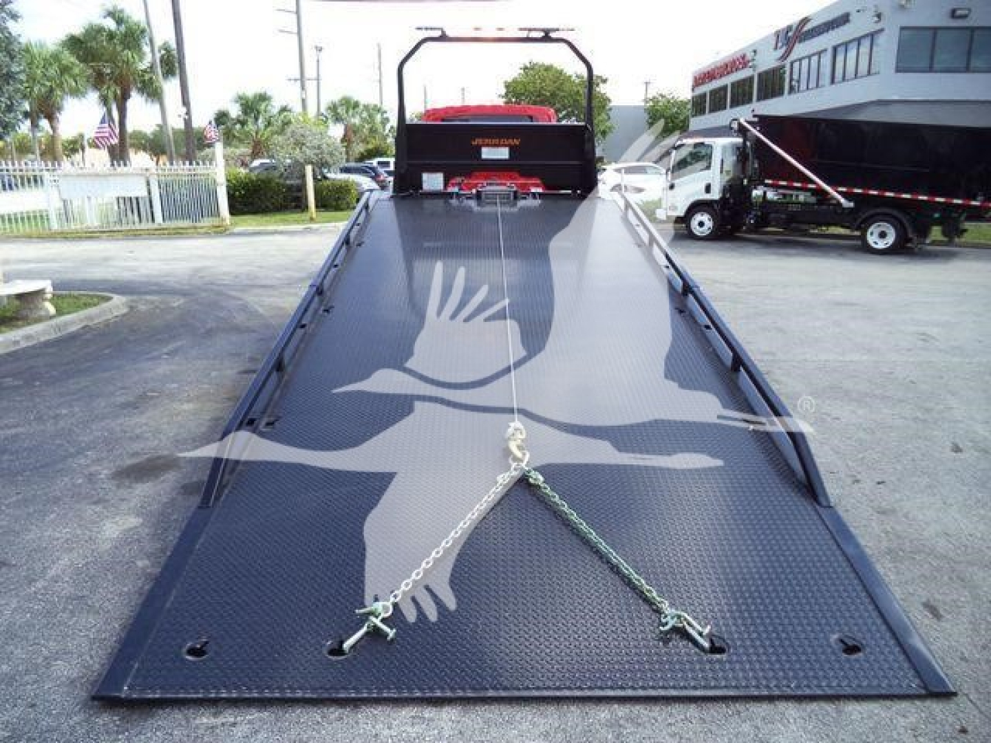Truck rollback equipment with a long black steel ramp, safety chains at the bottom, and a rear hitch, seen in a dealership lot.