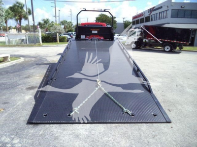 Flatbed tow truck deck with metal surface and hand crane at rear, chain lanyards across center, International MV stock unit in background.