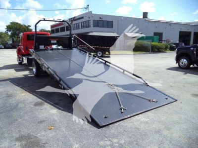 Truck rollback bed with hydraulic low-angle ramp extended, red tow truck cab in background, industrial building and other trucks visible.