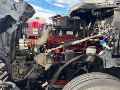 Mack PR64T76H engine bay showing red engine block, pink coolant reservoir, various hoses, wiring harness, air intake, and front wheel well.