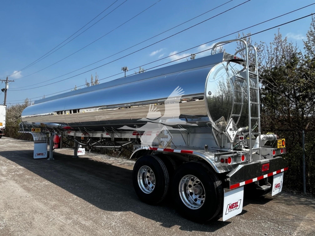 Large stainless steel Heil 9500 gallon tanker trailer with dual rear axles and polished rinse tank, reflective surface, safety rails, and rear bumper.