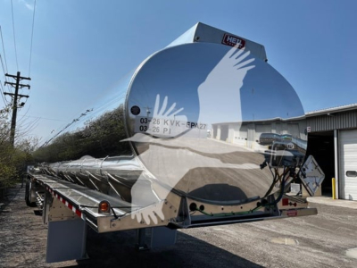 Rear view of a new Heil 4-Comp trailer showing the polished cylindrical tank with reflection of a person and surroundings; appears to be a tanker trailer with support frame and wheel fenders visible.