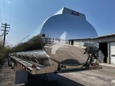 Heil 9500 gallon tanker trailer with polished stainless steel cylindrical tank, rear loading area, and mounted controls; reflective surface shows surrounding buildings.
