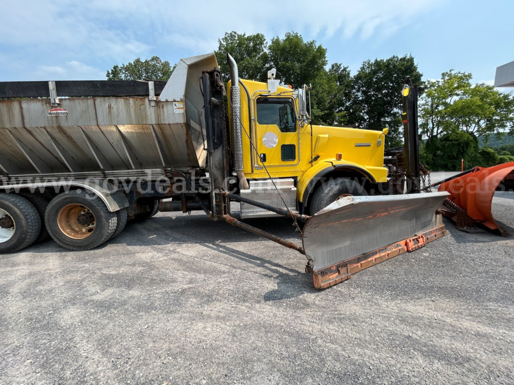 Kenworth T800 plow truck in yellow with a large snow plow blade attached, side view showing cab, front wheel, and dump bed partially visible.