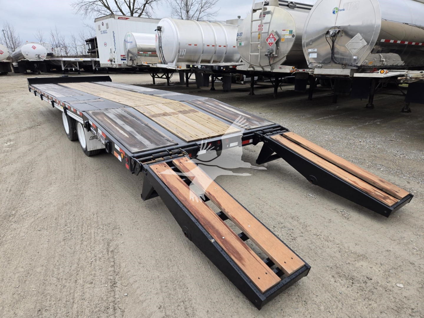 Trailer bed showing dual folding ramps and wooden deck with metal frame, in a yard with tanker trailers in background.