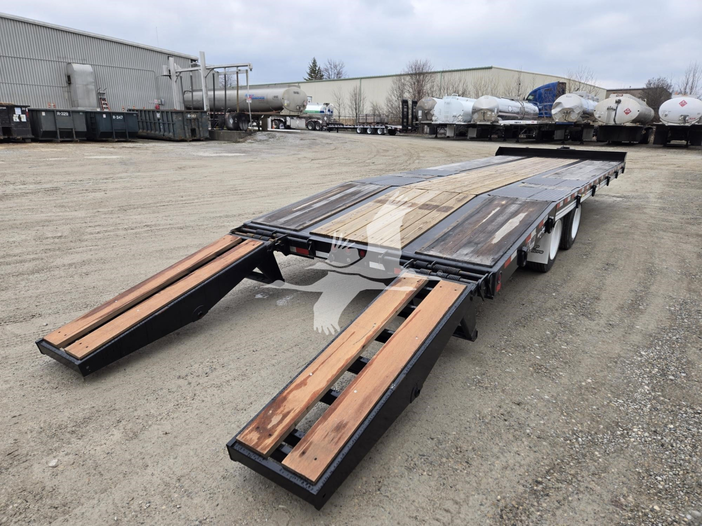 A long flatbed trailer with wooden planks on a two-axle gooseneck setup, in a truck yard with other trailers in the background.