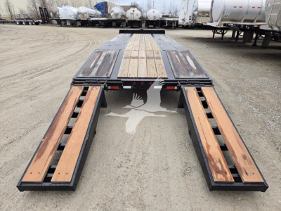 Flatbed trailer with wooden deck and side ramps in a dealership lot showing new trailer components and fuel tanks in background.