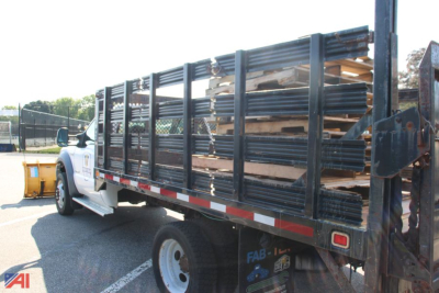 Flatbed truck with black stake bed rails loaded with lumber and planks, side view showing cab and rear plow hitch area.