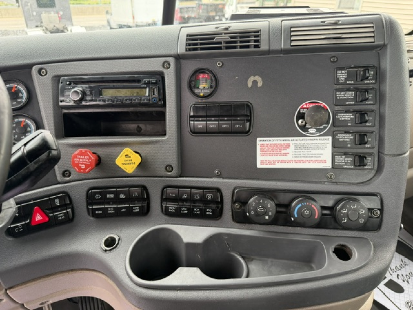 Dashboard of freightliner cascadia cab showing steering wheel, climate controls, radio, warning labels, and storage cup holders.