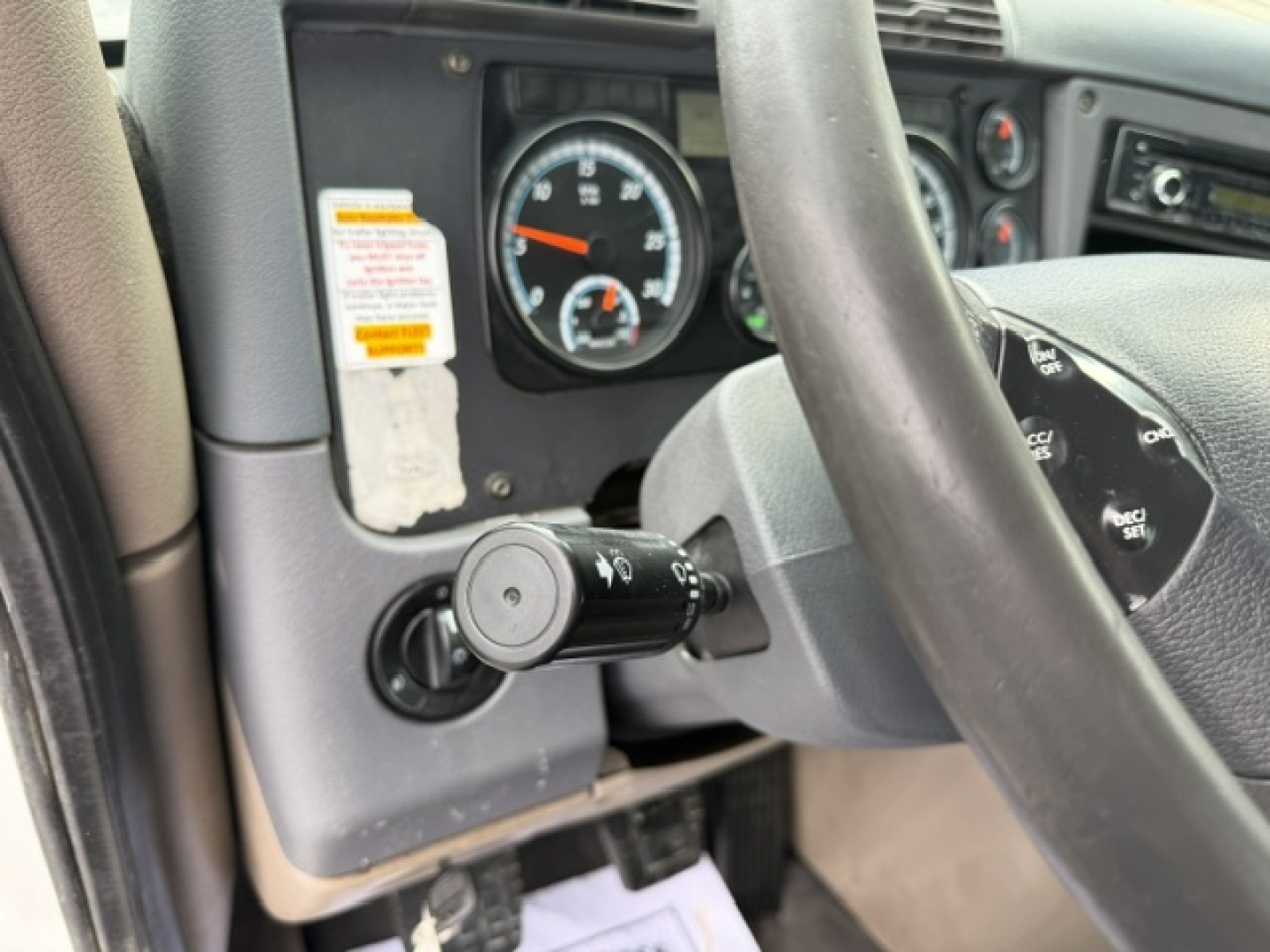Dashboard and steering column of a Freightliner Cascadia conventional daycab cockpit with tachometer, stalk controls, and steering wheel in view.