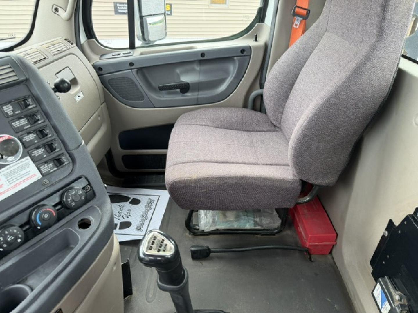 Cab interior of a Freightliner Cascadia daycab showing gray fabric seat and dash area with gear shifter and door panel