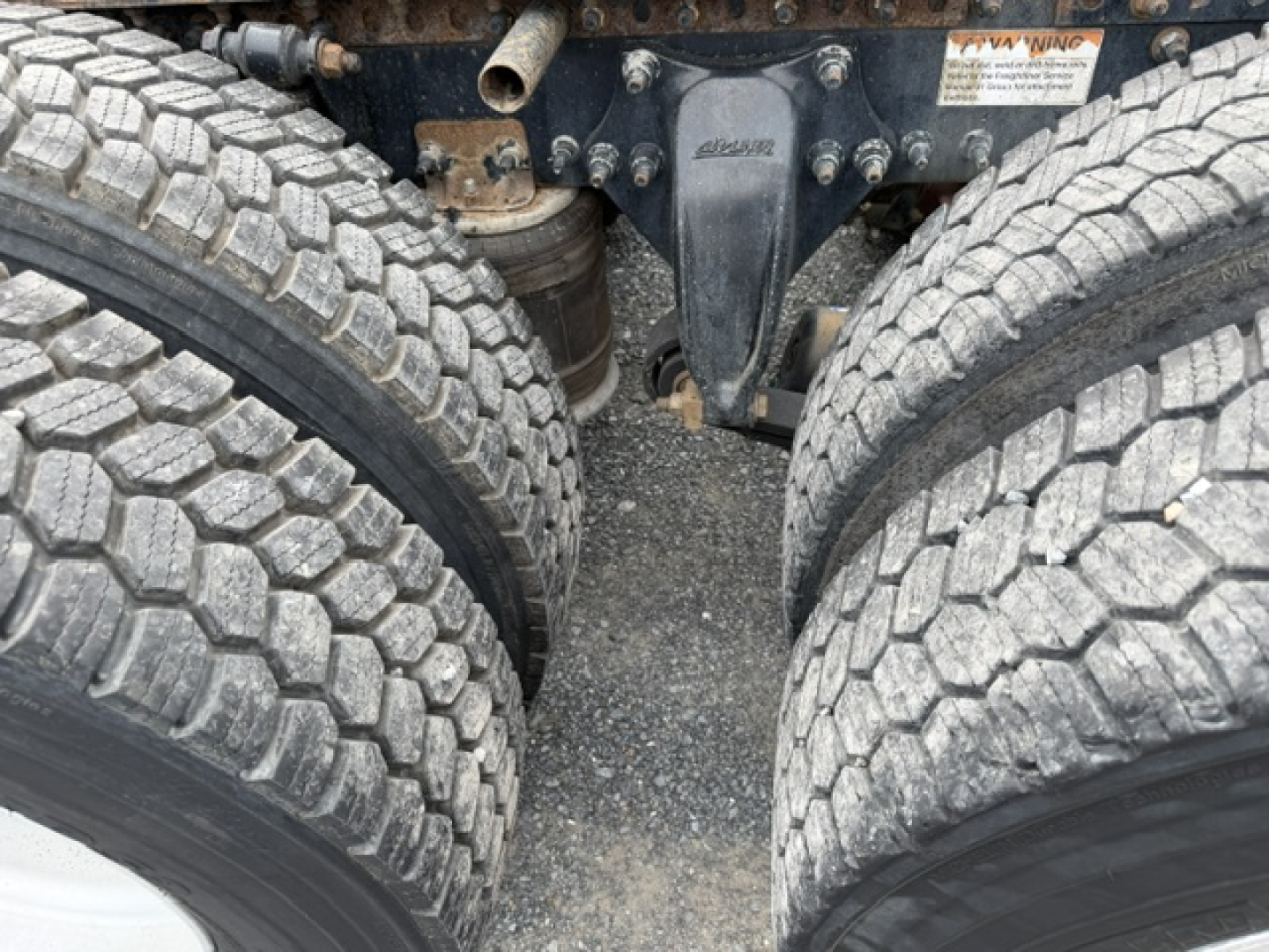 Rear view of Freightliner Cascadia chassis showing dual rear axles and suspension components; visible label with warning text on frame nearby.