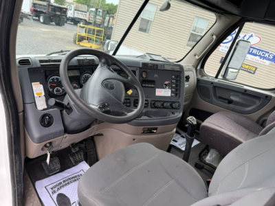 Freightliner Cascadia truck interior showing steering wheel, dashboard, and driver seat with gear shifter; monochrome gray-canvas seating and center display panel.