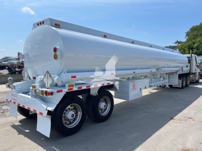 Tank trailer with cylindrical white tank on a dual-axle trailer. Visible text includes warning placards and reflective tape along the sides.