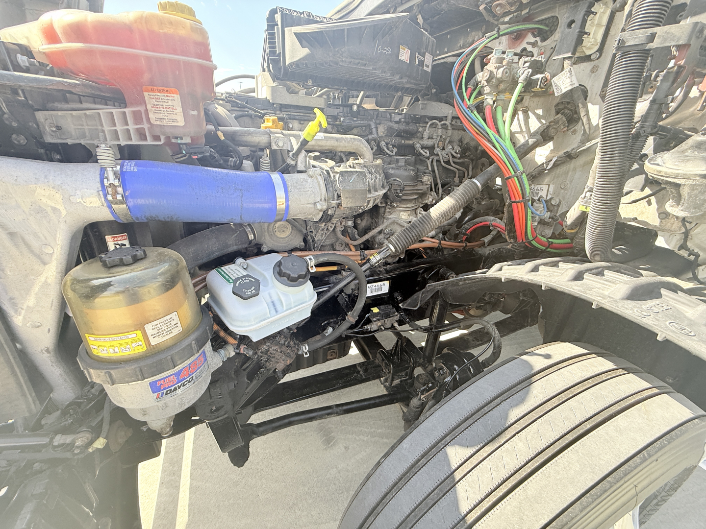 Truck engine bay showing power steering reservoir, brake fluid reservoir, blue silicone hose, and wiring harnesses in Freightliner PT126064S daycab.