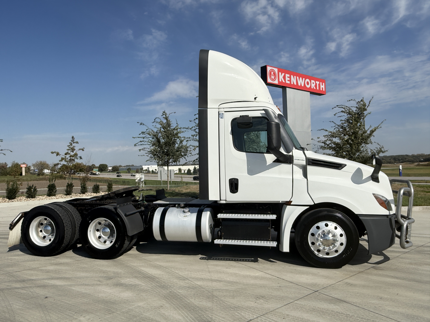 Truck side profile showing a white 2021 Freightliner PT126064S T conventional daycab with dual rear axles, chrome wheels, fuel tanks, and Kenworth sign in background.