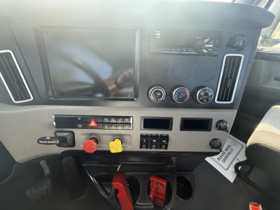 Cab dashboard of a Freightliner truck showing large infotainment screen, circular gauges, climate controls, two red/yellow toggles, and pedal area visible.