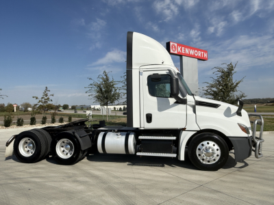 Truck side profile showing a white 2021 Freightliner PT126064S T conventional daycab with dual rear axles, chrome wheels, fuel tanks, and Kenworth sign in background.