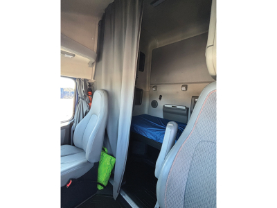 Truck cab sleeper area with partition curtain, captain seat in front, gray fabric seats, small bunk area behind curtain, interior walls and storage panels visible.