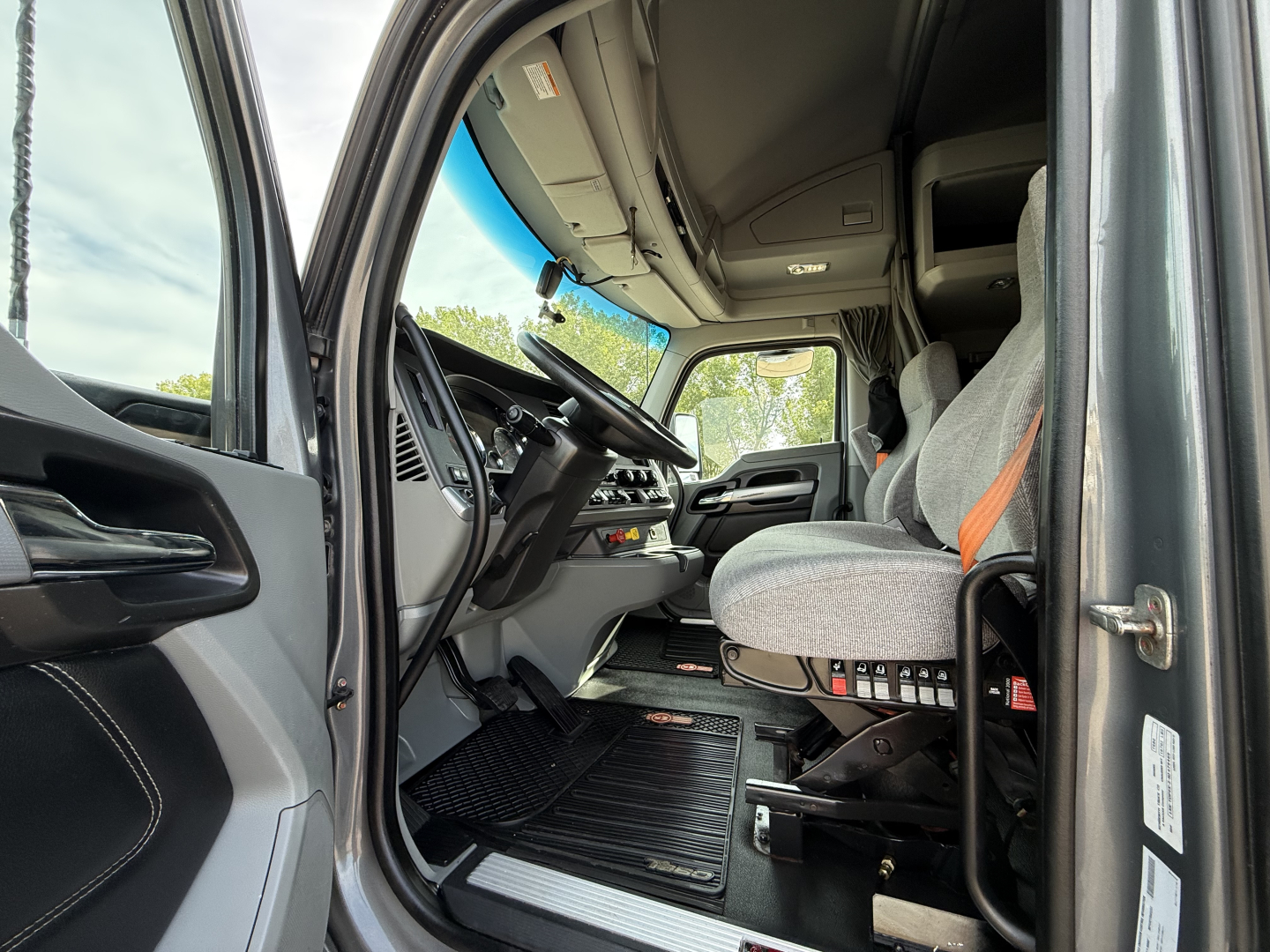 Kenworth T680 truck interior driver seat and dashboard view from open door showing steering wheel, pedals, and gray fabric seat.