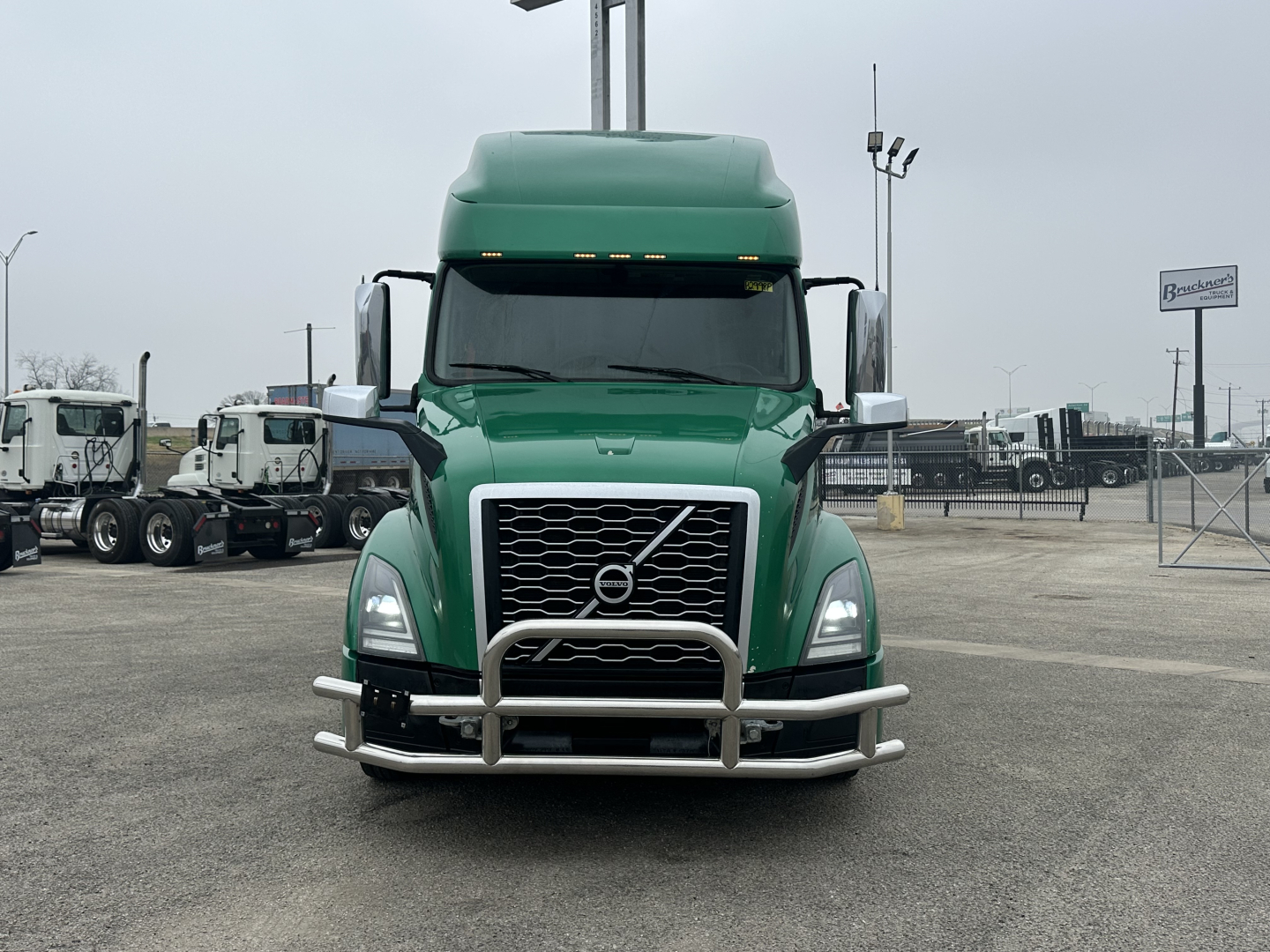 Volvo VNL64T760 truck front view showing large grille, chrome bull bar, green cab, and twin mirrors in an equipment lot.