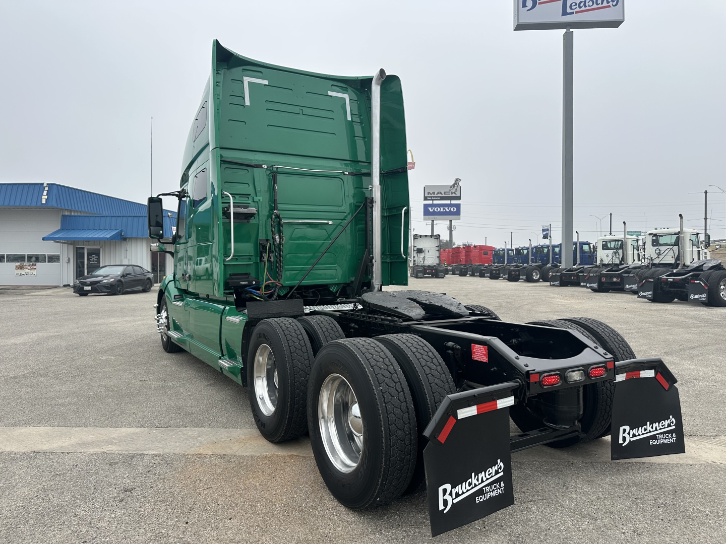 Truck rear and cab of a green Volvo VNL64T760 tractor. Twin rear axles, fifth wheel area visible, mud flaps with Bruchners logo, truck yard in background.