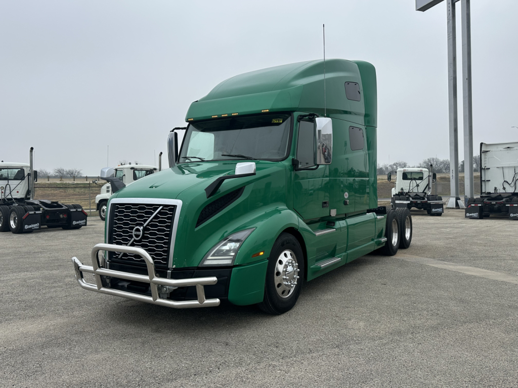 Green 2021 Volvo VNL64T760 sleeper tractor in a parking lot, front three-quarter view, with chrome bumper guard and grille.