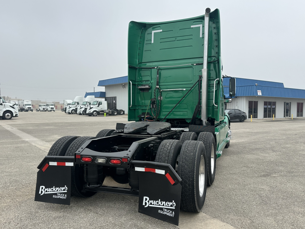 Rear view of a green Volvo VNL truck tractor with sleeper cab, dual rear axles, mud flaps, and fifth wheel missing visible; stock and dealership lot in background.
