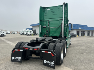 Rear view of a green Volvo VNL truck tractor with sleeper cab, dual rear axles, mud flaps, and fifth wheel missing visible; stock and dealership lot in background.