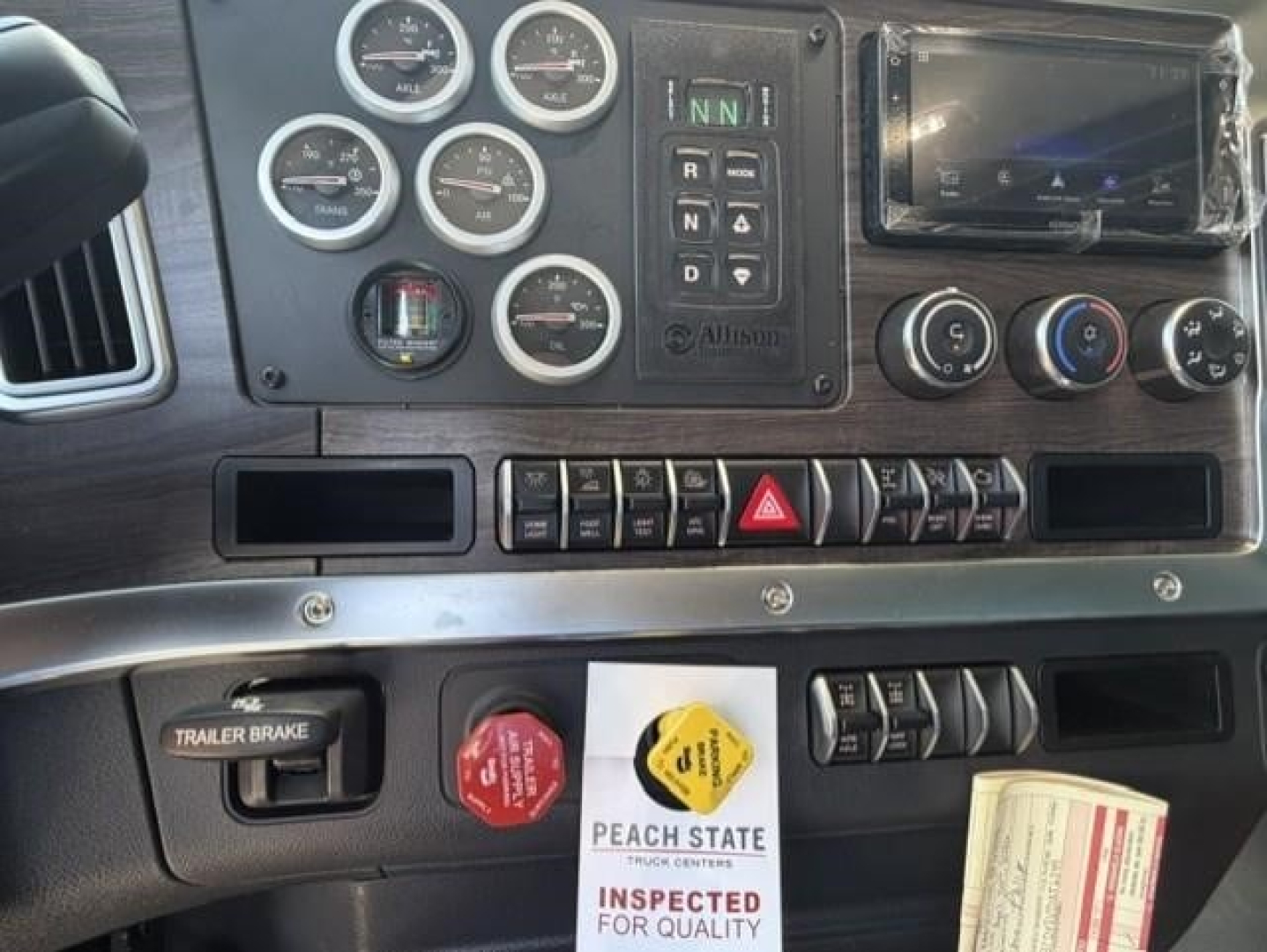 Dashboard panel of a new Western Star 47X truck showing multiple gauges, transmission gear selector, infotainment screen, hazard switch, trailer brake, and inspection tag.