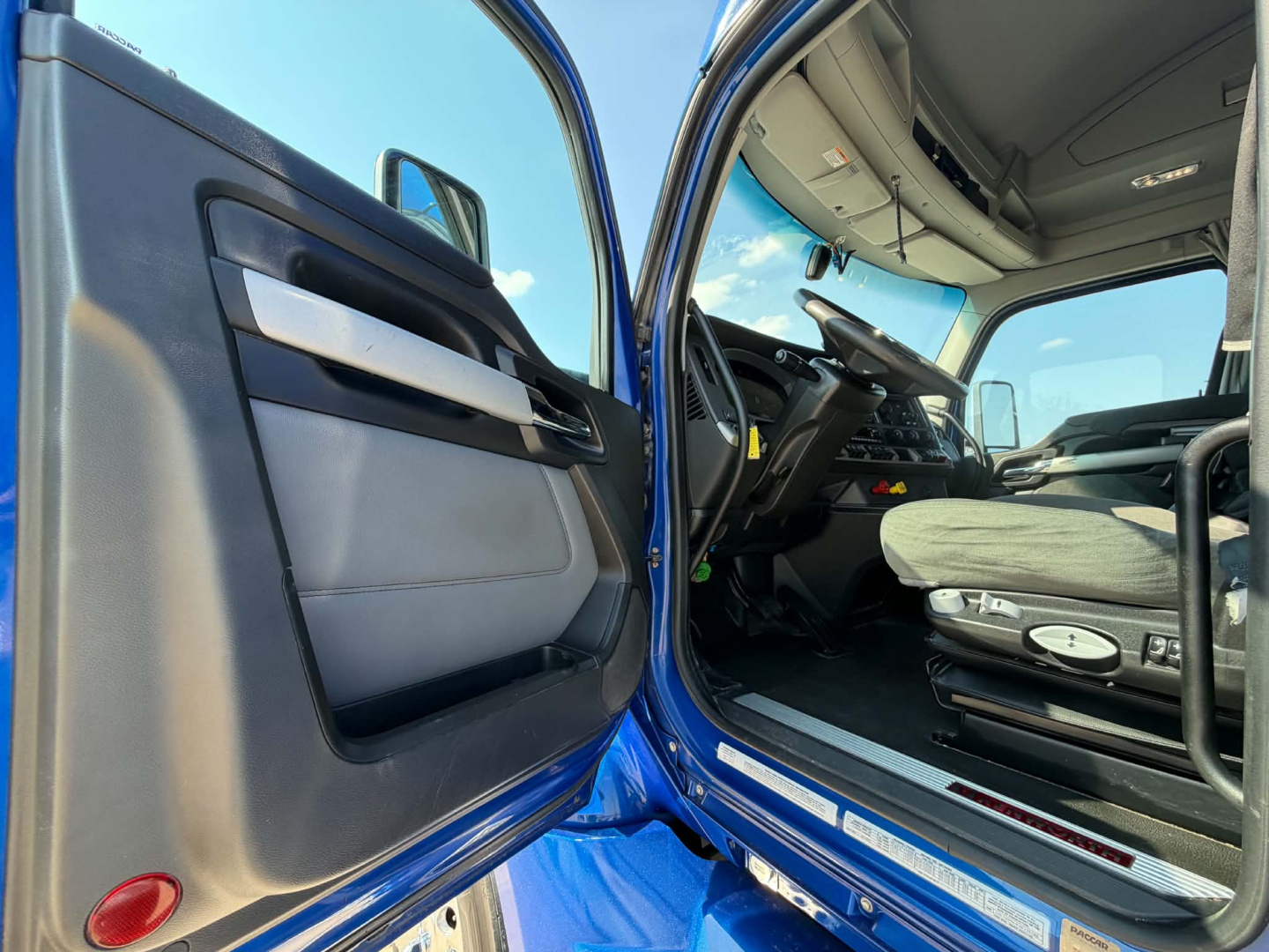 Truck cab interior showing open driver door with blue exterior, steering wheel, driver's seat, and dashboard view.