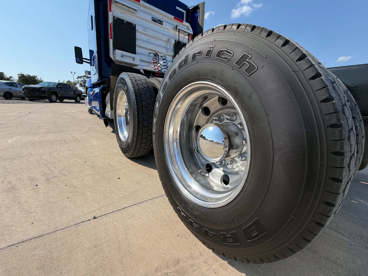 Front-right wheel and dual rear tires of a blue 2022 Kenworth T680 tractor-trailer showing chrome hub, Bridgestone tires, and sleeper cab in background.