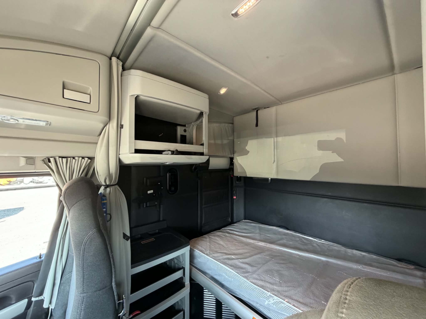 Interior of a 2022 Kenworth T680 sleeper cab showing bunk bed with mattress, overhead storage cabinets, and a small shelf with built-in storage.