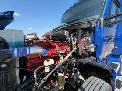 Kenworth T680 engine bay showing red V6 or V8 engine with exposed components, hoses, wiring, reservoir, and blue cab; various fluid lines and intake components visible.