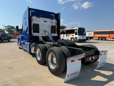 Kenworth T680 rear-angled view showing dual rear axles with mud flaps and fifth-wheel area, blue cab, chrome wheels, and fuel/air lines.