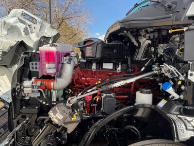 Engine bay of a new 2026 Mack PR64T76M showing red engine, pink coolant reservoir, black hoses, wiring, and metal components.