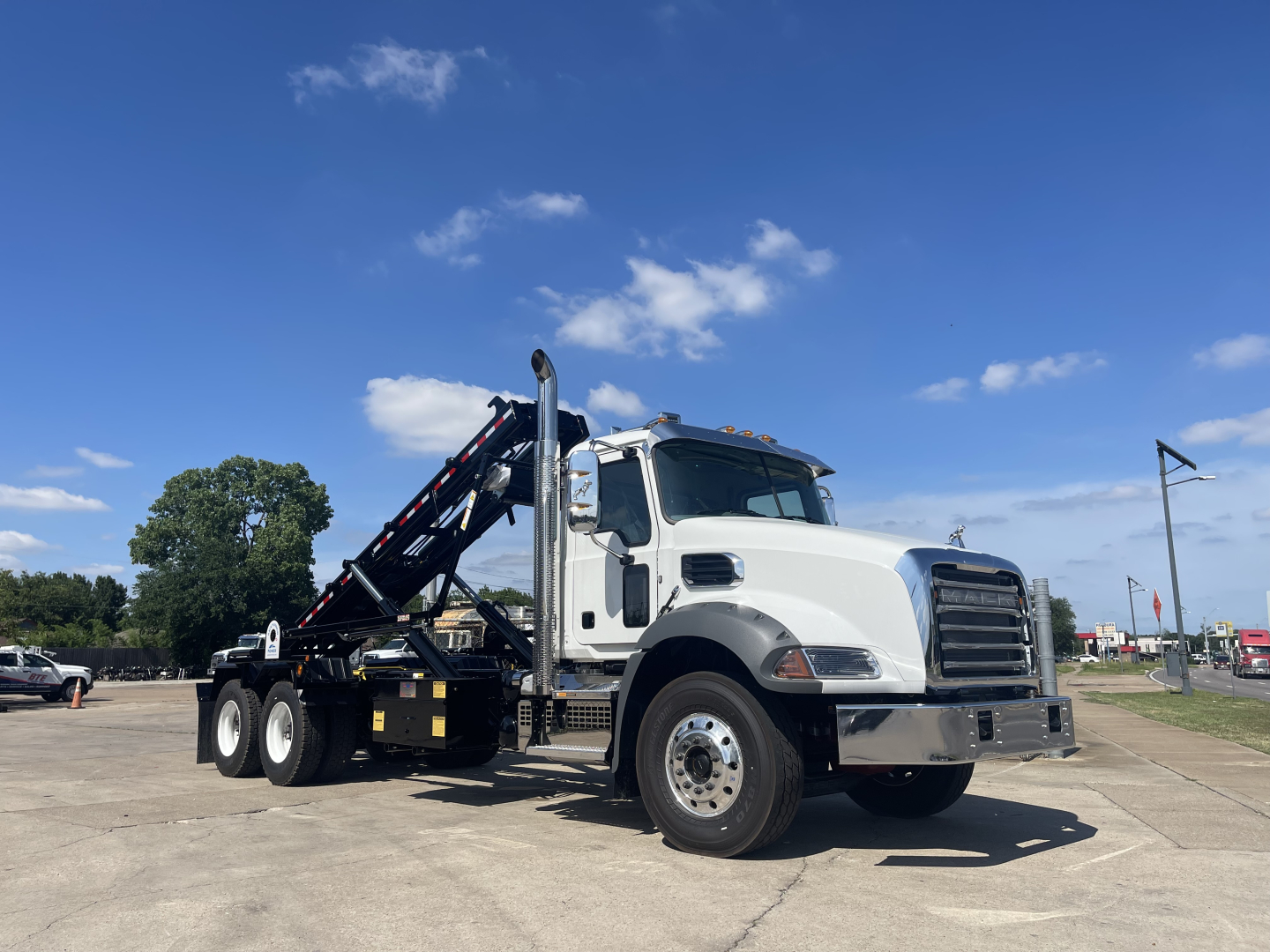 Truck cab in front of a roll-off container system, 2027 Mack GR64B, white cab, black hydraulic arm, three rear axles, side view with blue sky