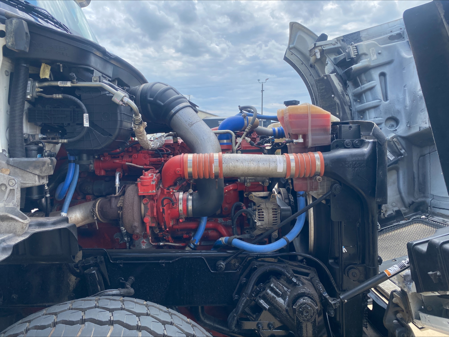 Kenworth T880 engine bay view showing red engine, blue and orange hoses, air intake, and radiator with coolant reservoir cap.