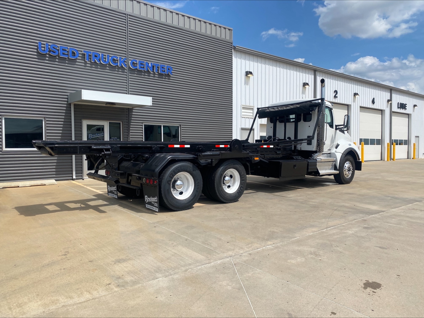 2019 Kenworth T880 roll-off truck with long flatbed and rear wheel configuration visible. Text on building reads USED TRUCK CENTER. VIN sticker and stock number not clearly readable on image.