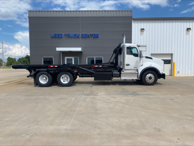 Kenworth T880 roll-off truck side view showing white cab and black long flatbed with hydraulic equipment and three rear axles.