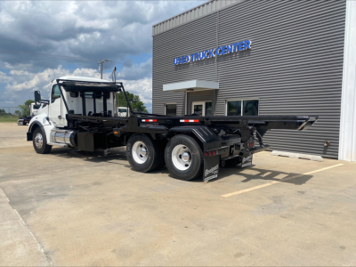 Kenworth T880 roll-off truck in front of used truck center with flatbed/haul trailer and multiple wheels visible.