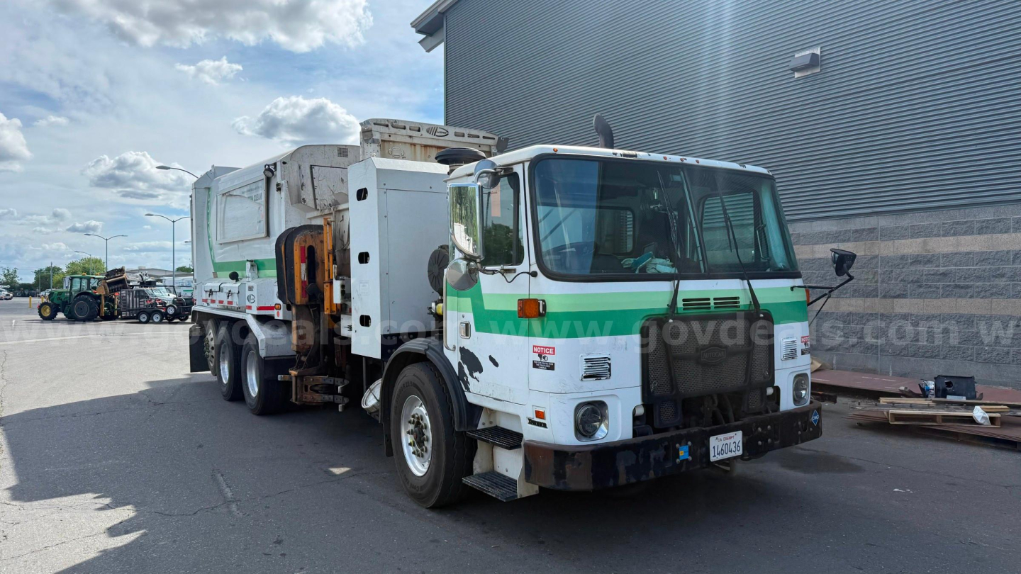 Autocar Xpeditor garbage truck in outdoor lot, front view showing cab, green and white livery, garbage body, warning lights on top, side mirrors, heavy-duty bumper.