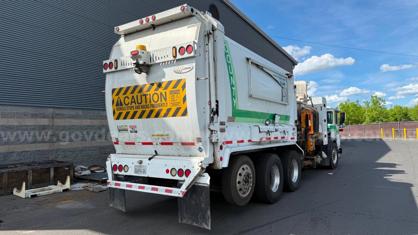 Rear and side view of a white Autocar XPeditor garbage truck with a large hopper, safety CAUTION stripe, and multiple rear lights.