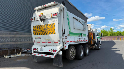 Rear and side view of a white Autocar XPeditor garbage truck with a large hopper, safety CAUTION stripe, and multiple rear lights.
