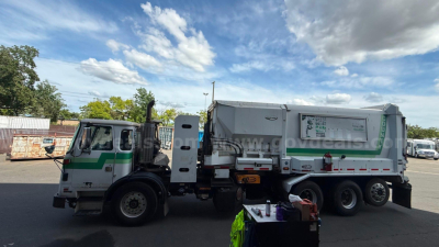 Autocar Xpeditor truck with a green and white cab and a large waste collection body; side view shows a rear cab access door and hydraulic components.