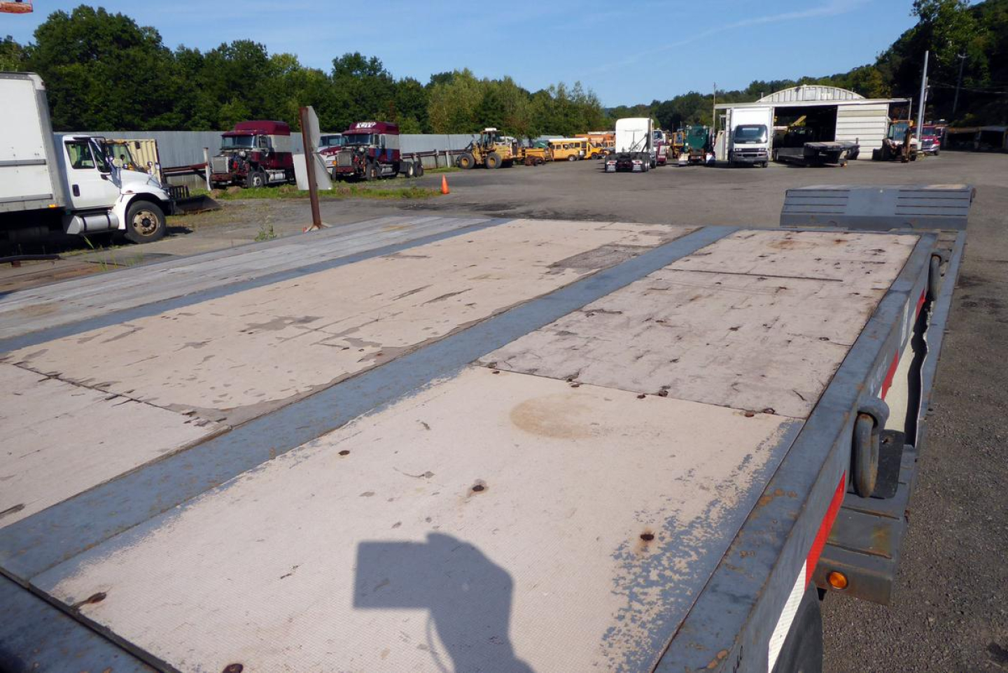 Flatbed trailer deck with plywood surface and steel edging seen from the side, industrial yard in background.
