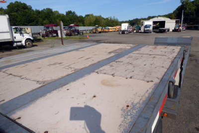 Flatbed trailer deck with plywood surface and steel edging seen from the side, industrial yard in background.