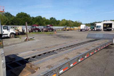 Trailer bed with lowered trestle-style ramp sections in a truck yard; other trucks and trailers in background under a clear blue sky.