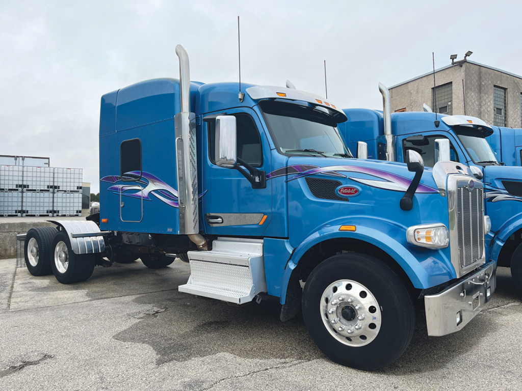 Peterbilt 567 UltraCab truck in blue with chrome exhaust stacks and graphics on the door, side view showing front wheel, fender, and grille.